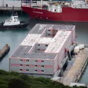 A tug boat passes the Bibby Stockholm accommodation barge at Portland Port in Dorset