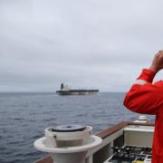 Image shows a man in a US Coast Guard jacket looking at a ship through binoculars